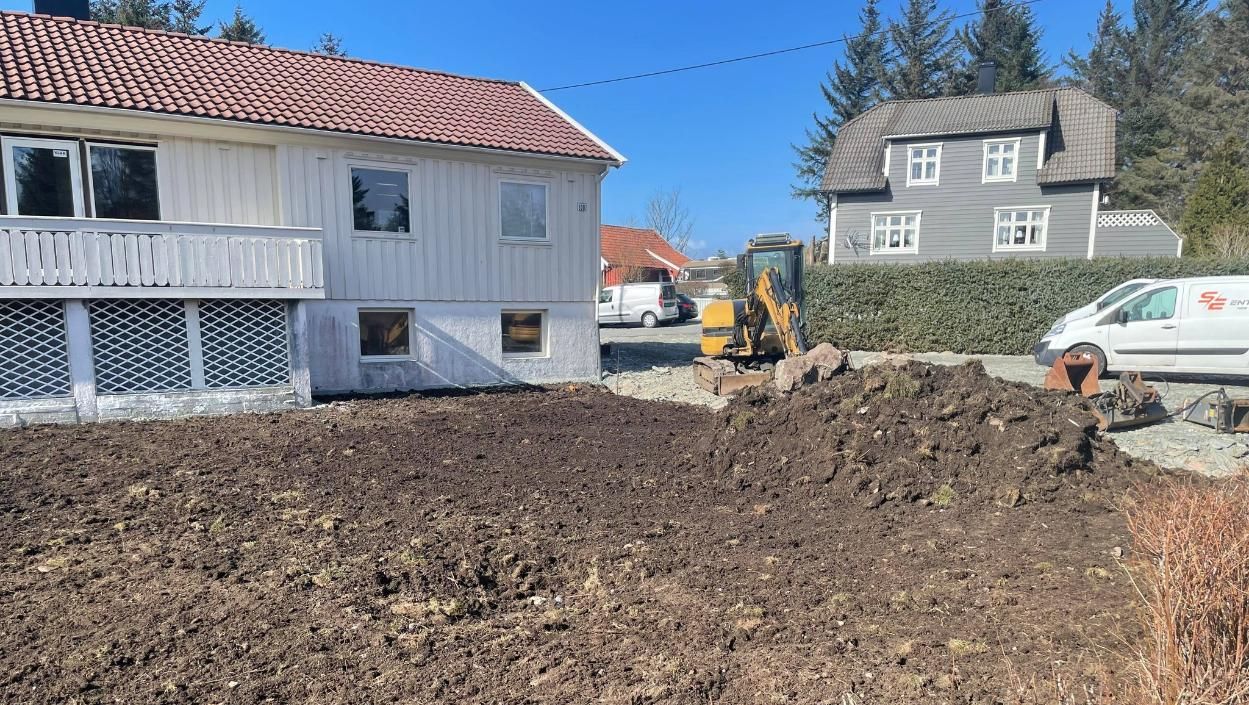 Enebolig med hvite vinduskarmer og terrasse, med et nypløyet jordstykke foran. En liten gravemaskin jobber til høyre i bildet, med en grå husfasade i bakgrunnen.