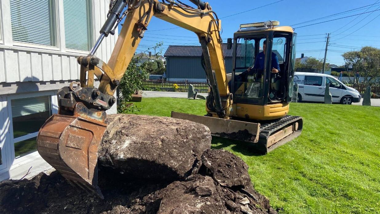 Gravemaskin løfter stor stein ved husets grunnmur